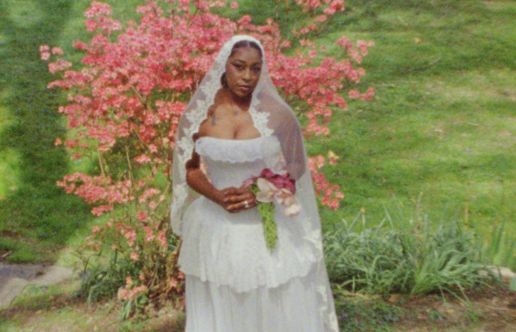 The bride in a lacey corseted wedding gown and mantilla veil posing behind a pink cherry blossom tree in Asheville, North Carolina. Captured on Super 8 film
