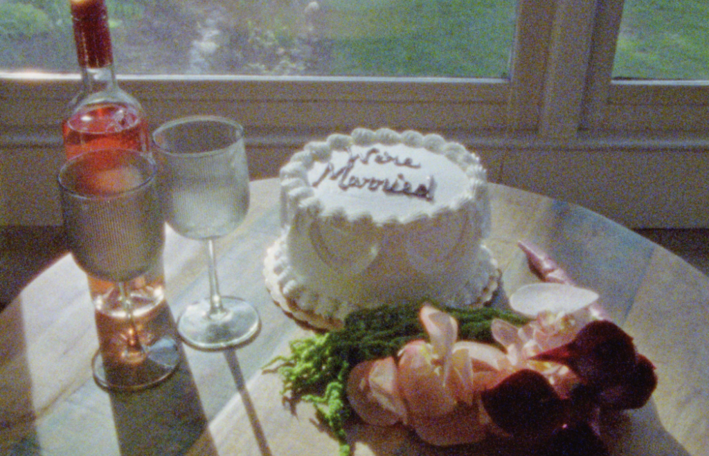 A Super 8mm Still frame of the inside of an Asheville Airbnb dining table with a white vintage wedding cake, pink orchid bouquet, and rose wine. The white vintage cake says "We're Married," as the couple is celebrating their elopement.