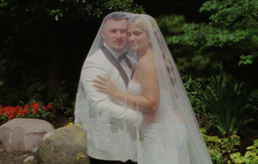 A Super 8mm still frame of the bride and groom embracing, a lace cathedral veil draped over them, inside a garden in Plainfield, IL