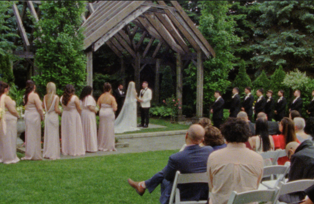 A wide shot on Super 8mm film of the bride and groom at the alter at the outdoor chapel, with bridesmaids, groomsmen, and guests surrounding them during their ceremony