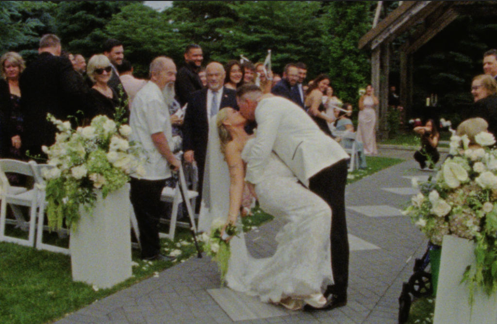 A Super 8mm film still frame of the bride and groom dipping into a kiss as they exit the ceremony space in Plainfield, IL